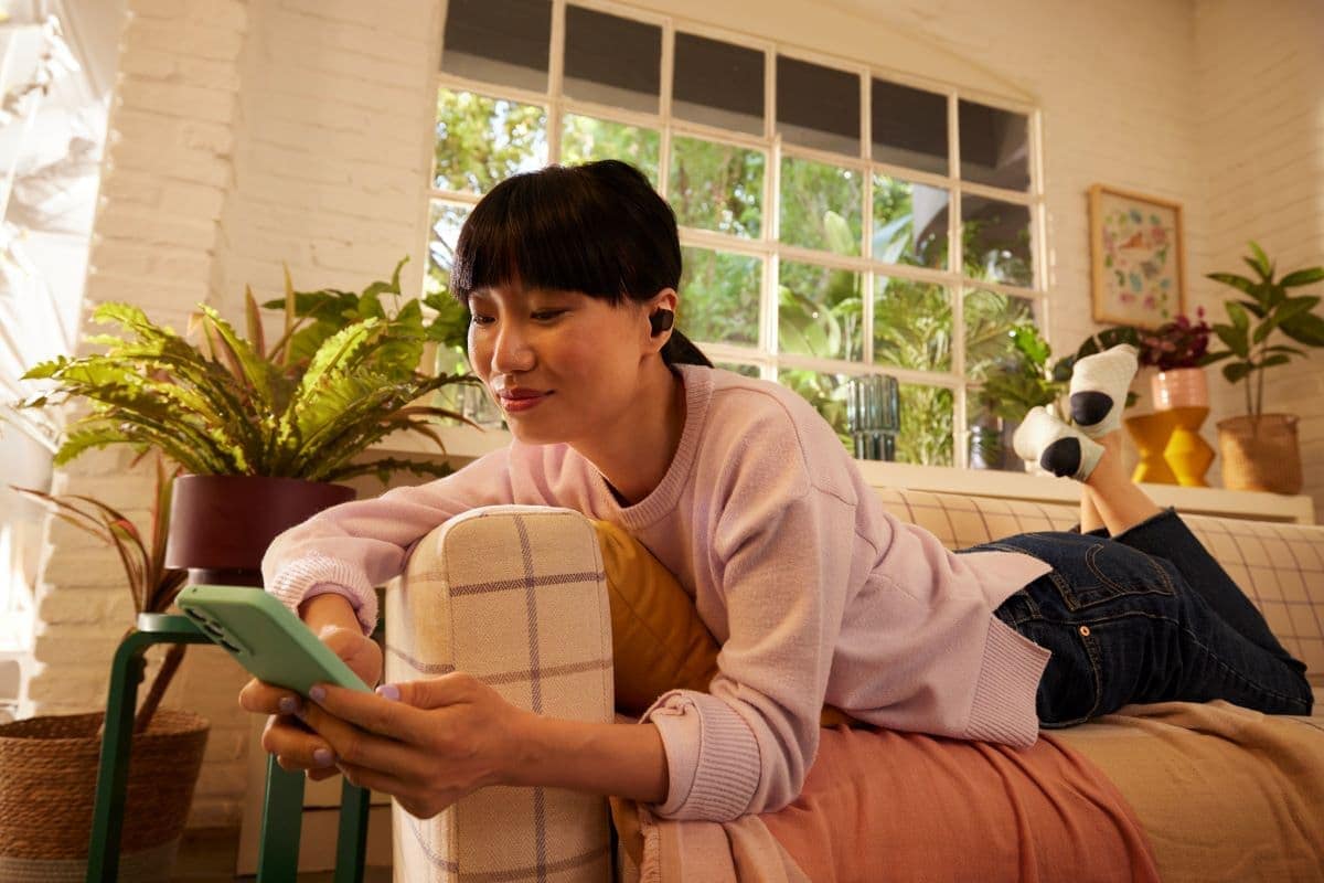 A young woman with short dark hair relaxes on a couch in a sunny room filled with plants, looking at her phone while wearing wireless poly voyager earbuds